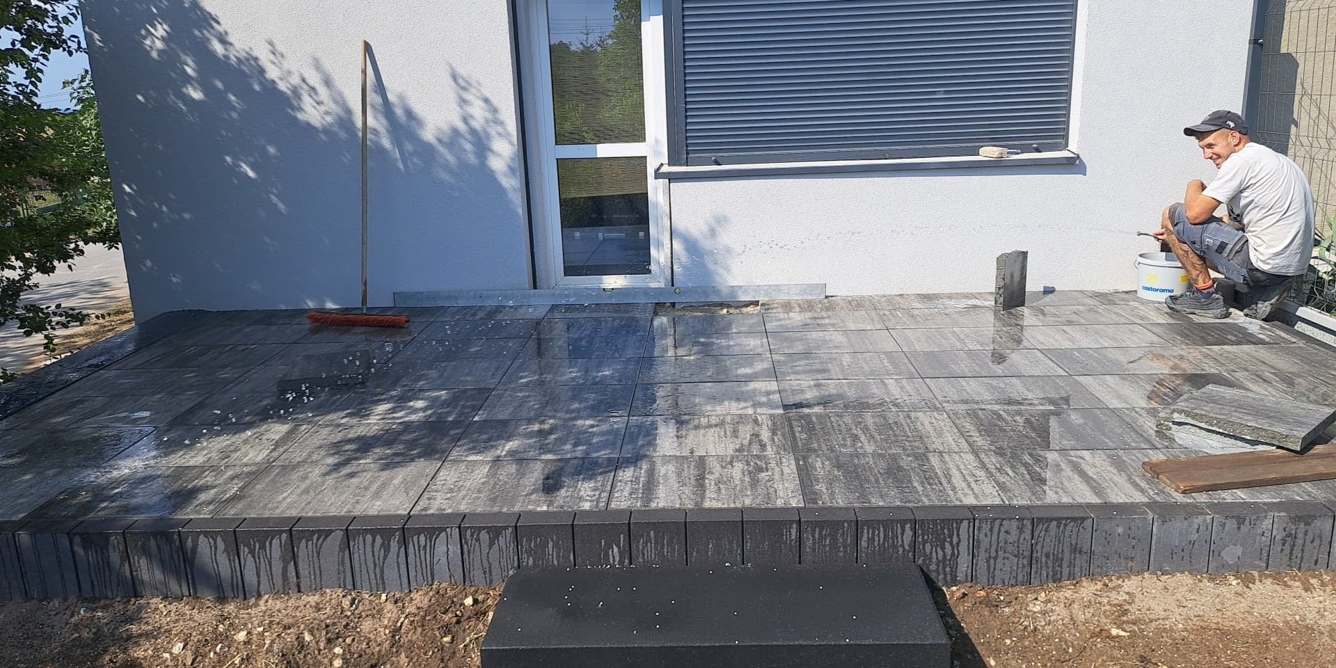 Pracownik w czapce czyści wodą świeżo ułożony taras z kostki brukowej w odcieniach szarości, z widoczną elewacją budynku i oknem z roletą.