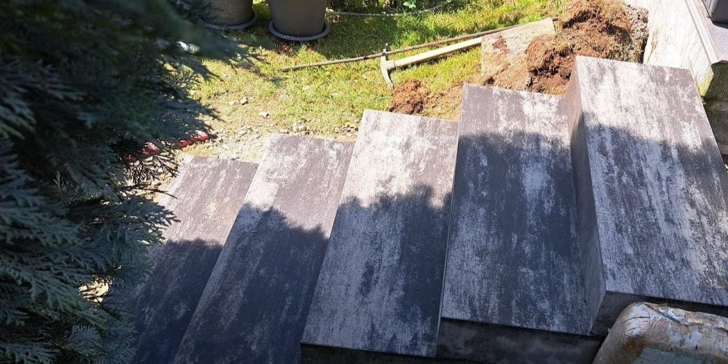 Schody zewnętrzne wyłożone szarą kostką granitową, widok z góry z fragmentem iglastego krzewu po lewej stronie i narzędziem ogrodniczym oraz ziemią w tle.