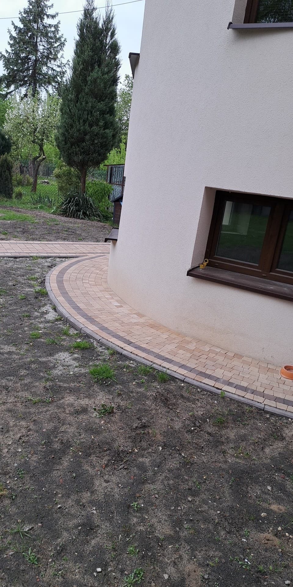 Zakrzywiony chodnik z kostki granitowej w odcieniach beżu i brązu, przylegający do zaokrąglonej ściany budynku z oknem. W tle widoczne drzewa i trawnik.