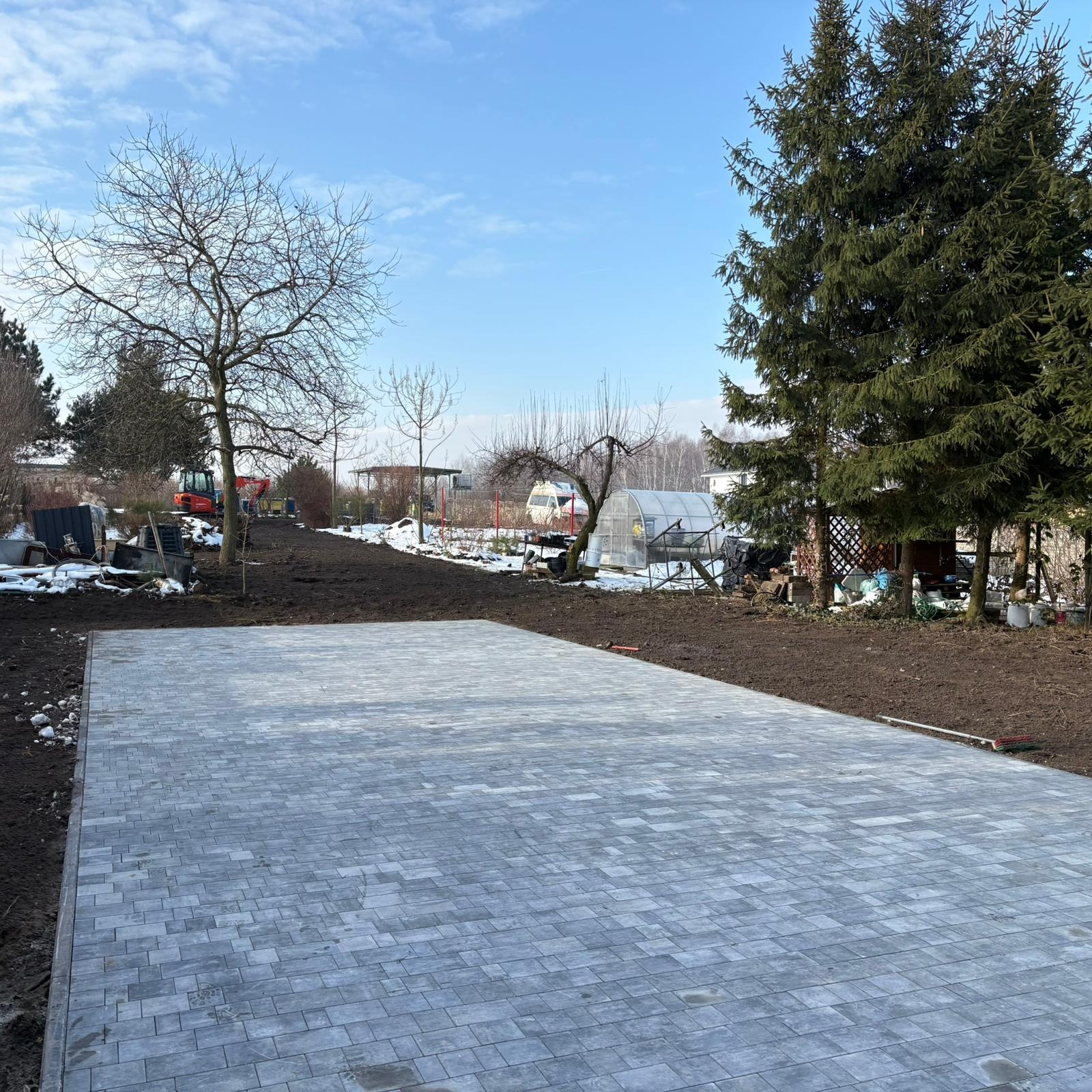 Ułożona szara kostka brukowa na posesji, w tle widoczny minikoparka i zabudowania gospodarcze, częściowo pokryte śniegiem