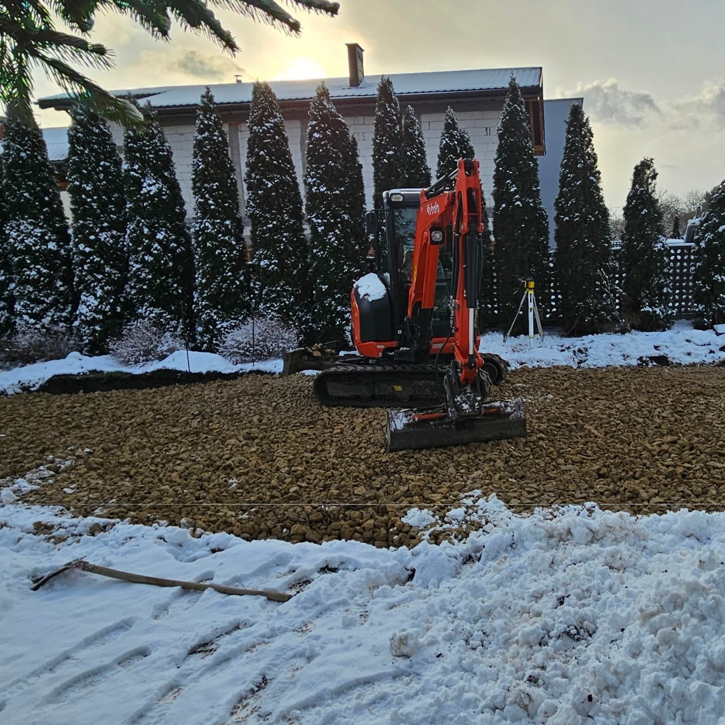 Pomarańczowa mini koparka Kubota na placu budowy w zimowej scenerii, teren przygotowany i wysypany kamieniami, w tle rząd wysokich, zimozielonych drzew i budynek.