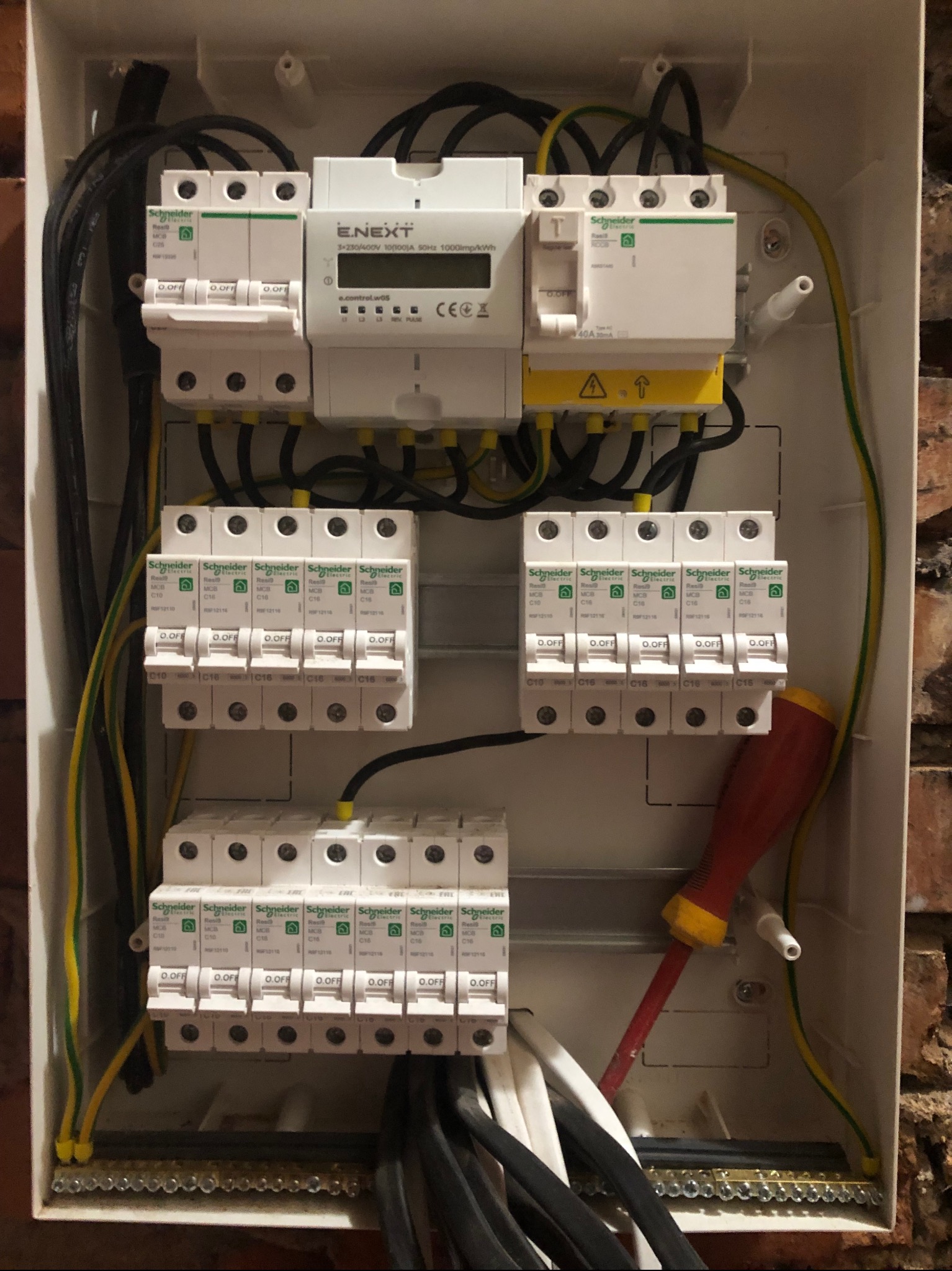 Rozdzielnia elektryczna z zamontowanymi bezpiecznikami i licznikiem, widoczne przewody i narzędzie (śrubokręt) wewnątrz obudowy. Schneider Electric.