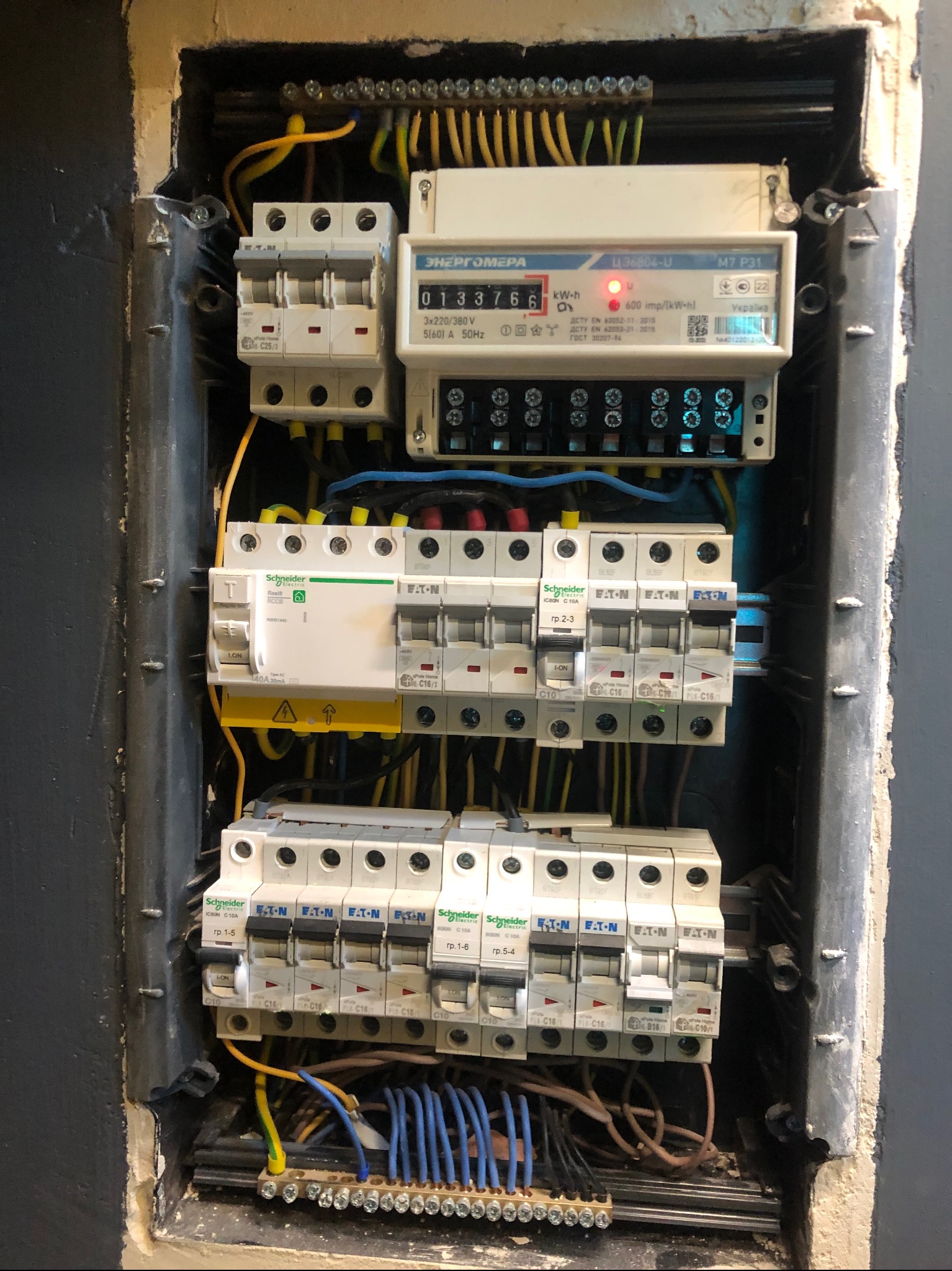 Rozdzielnia elektryczna z bezpiecznikami, licznikiem energii i okablowaniem w szarej obudowie. Widoczne elementy marki Schneider Electric i Eaton.