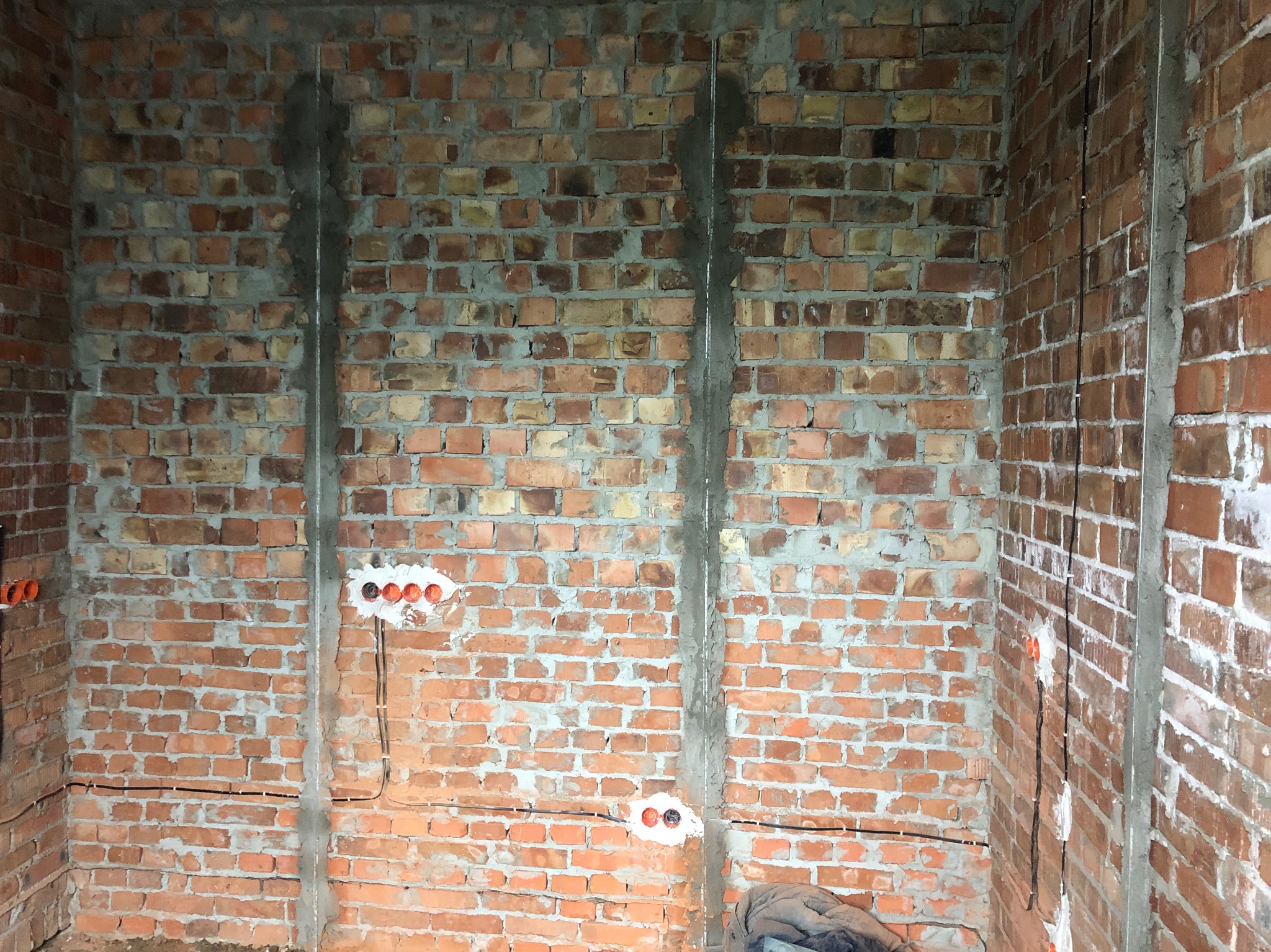Czerwona ceglana ściana z widocznymi przewodami elektrycznymi i puszkami. Instalacja w trakcie montażu, widoczne kable i wyżłobienia w ścianie.