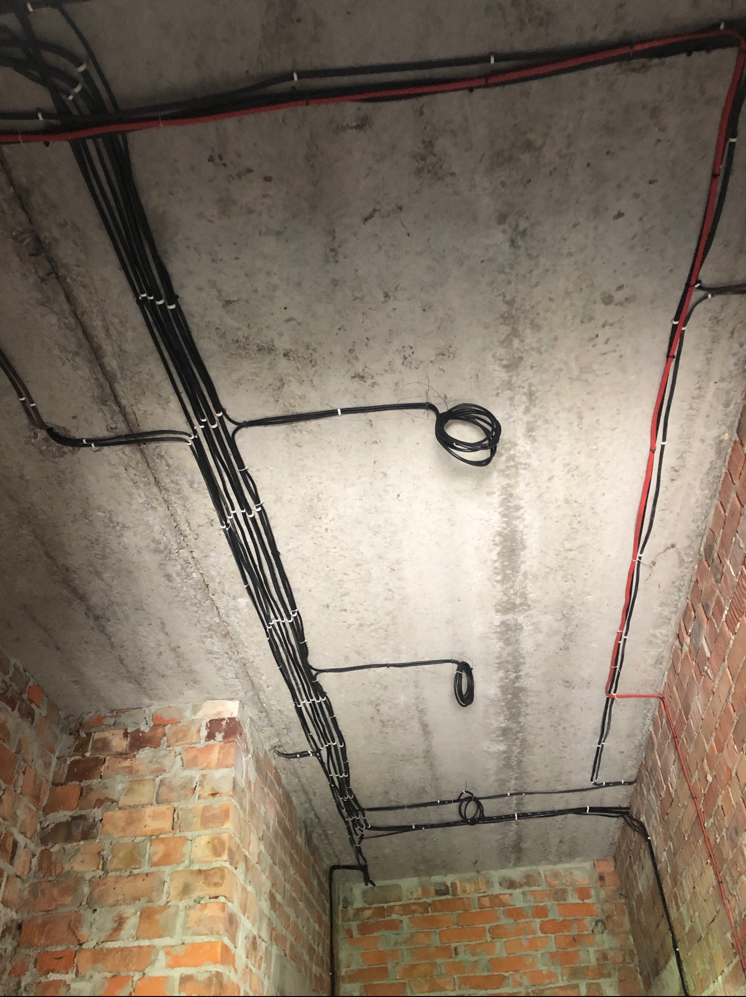 Surowy betonowy strop z instalacją kablową w Radziszewie. Kable czarne i czerwone, mocowane uchwytami. Część kabli zwinięta w pętle. Ściany z czerwonej cegły.