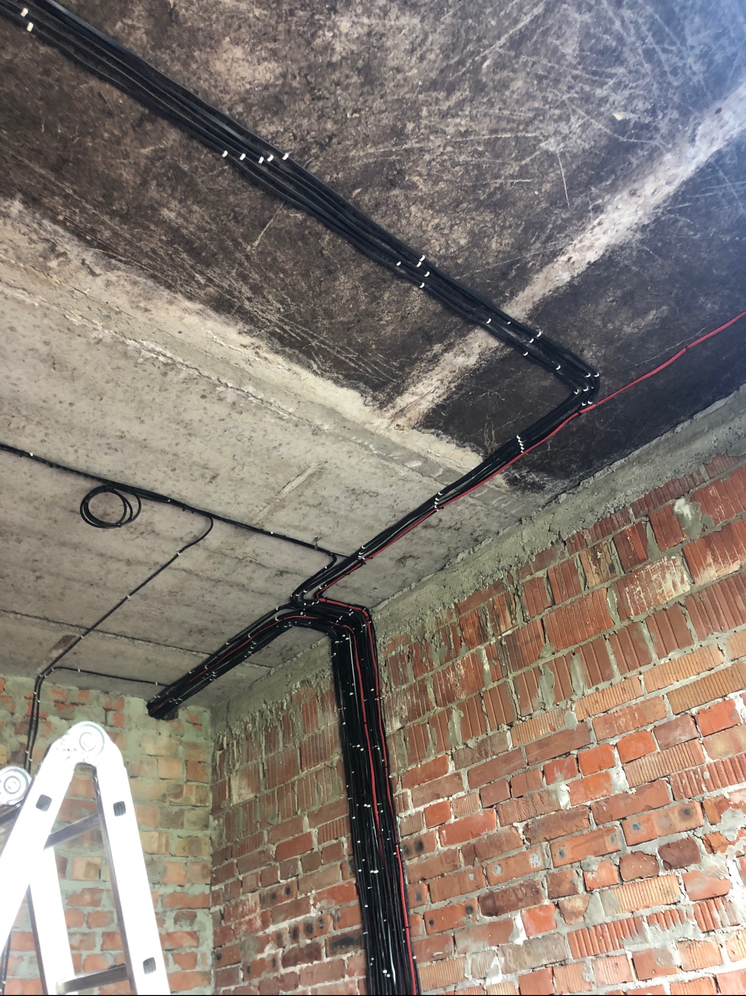 Czarno-czerwone kable elektryczne prowadzone po suficie i ceglanej ścianie w surowym stanie, widoczne mocowania, drabina w lewym dolnym rogu.