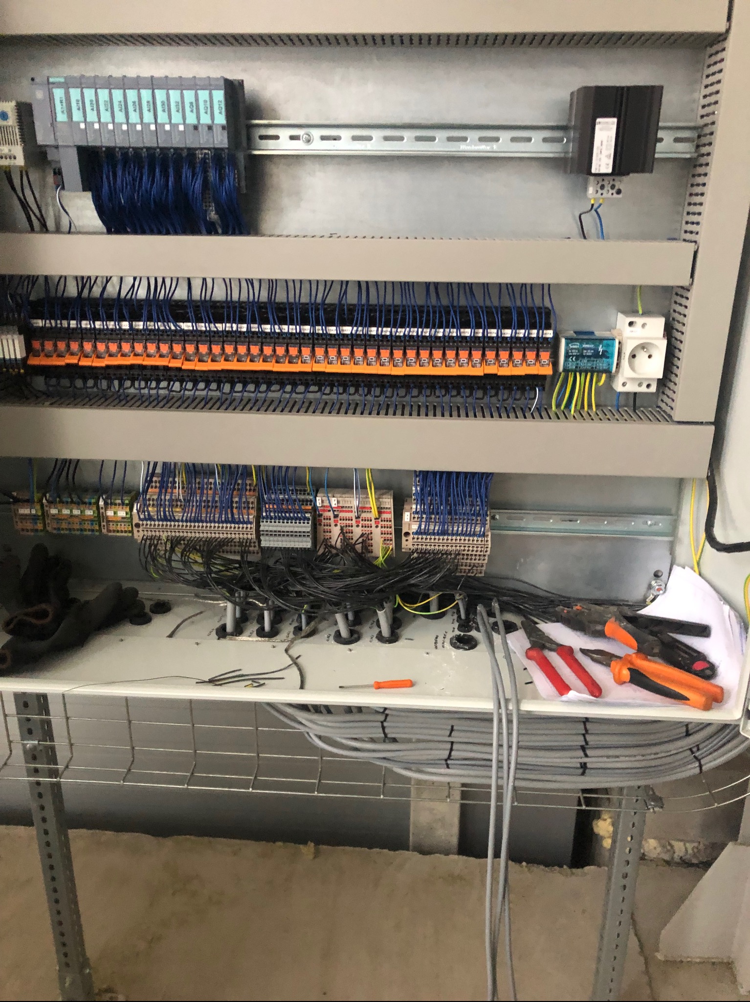 Rozdzielnica elektryczna w trakcie montażu, widoczne kable, listwy zaciskowe, bezpieczniki i narzędzia (szczypce, wkrętak). Częściowo widoczne rękawice robocze.