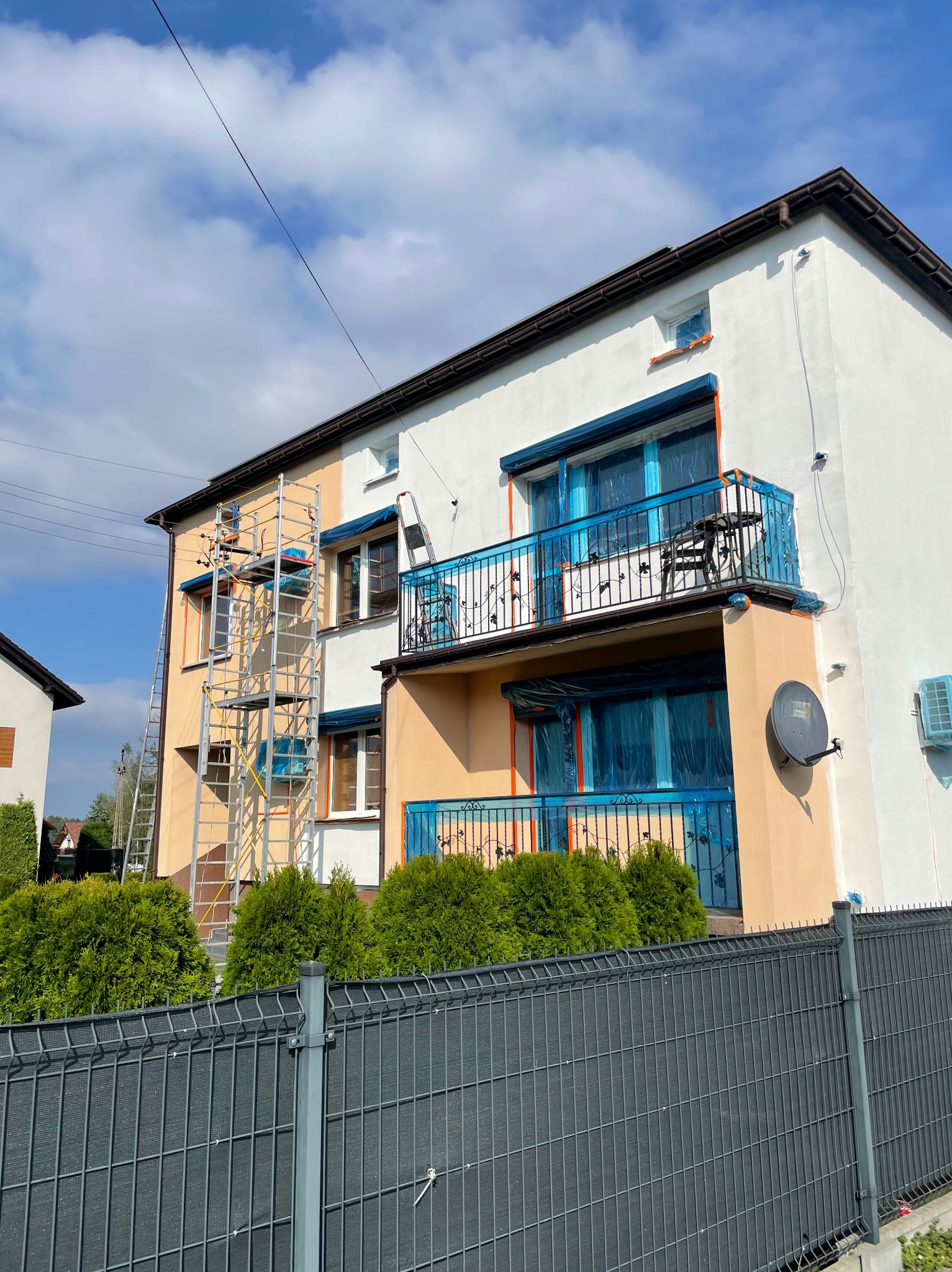 Dwukondygnacyjny dom w trakcie malowania elewacji. Część ściany pomalowana na biało, druga w kolorze beżowym. Okna zabezpieczone folią. Rozstawione rusztowania.