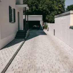 Ułożona, jasnoszara kostka brukowa na podjeździe z odpływem liniowym, prowadząca do garażu z zaparkowanym samochodem i schodów wejściowych do domu.