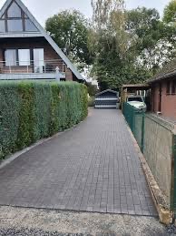 Ułożona, szara kostka brukowa prowadząca do garażu z białymi drzwiami, obok zadbany żywopłot i dom z balkonem.