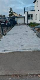 Świeżo ułożona, szara kostka brukowa na podjeździe przed domem, widok z poziomu chodnika, z widocznymi krawężnikami i ogrodzeniem w tle.