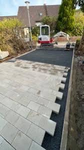 Fragment układanej nawierzchni z jasnoszarej kostki brukowej, widoczny mini-koparka w tle, przygotowane podłoże z czarnego grysu.