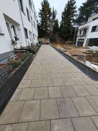 Długa, wyłożona jasnymi, kwadratowymi płytami ścieżka prowadząca wzdłuż białego budynku mieszkalnego, z obrzeżami z ciemnych kamieni, widok z perspektywy chodnika.