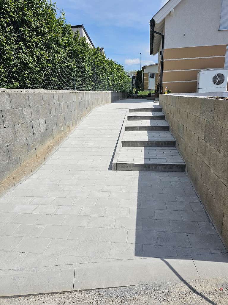 Podjazd i schody wyłożone szarą kostką brukową prowadzące do domu, otoczone murem z betonowych bloczków i zielonym żywopłotem.