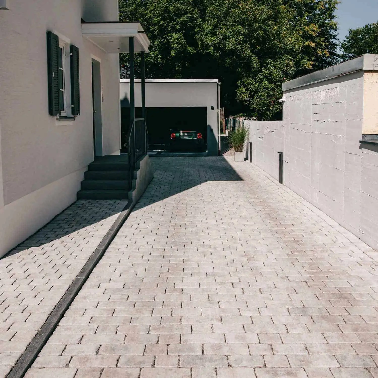 Ułożona, jasnoszara kostka brukowa na podjeździe z odpływem liniowym, prowadząca do garażu z zaparkowanym samochodem i schodów wejściowych do domu.