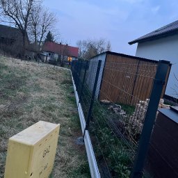 PETRO TOKARENKO - Nowoczesne ogrodzenie panelowe w kolorze grafitowym z podmurówką, widok z perspektywy skrzynki gazowej na tle zabudowań w pochmurny dzień.