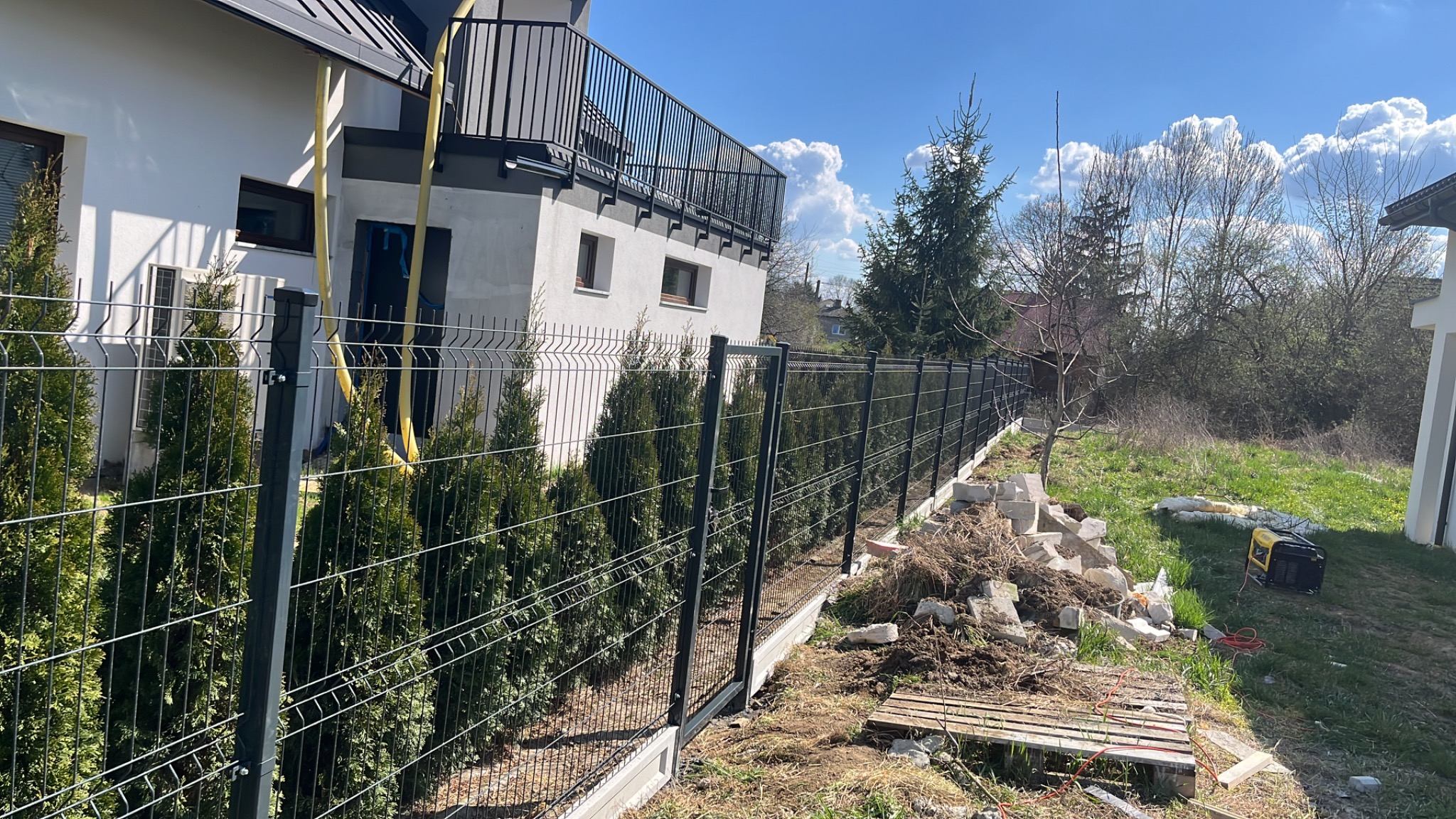 Nowoczesne ogrodzenie panelowe w kolorze antracytowym, z podmurówką betonową, oddzielające posesję z białym domem i balkonem od sąsiedniej działki z widocznymi drzewami i generatorem prądu.