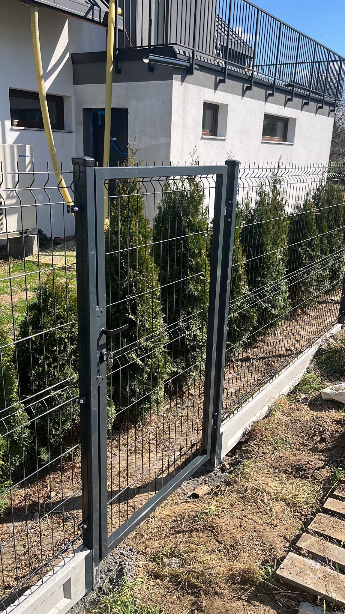 Nowoczesna, grafitowa furtka z siatki panelowej, z widocznymi kluczami w zamku, prowadząca do ogrodu z tujami, na tle białego domu z balkonem.