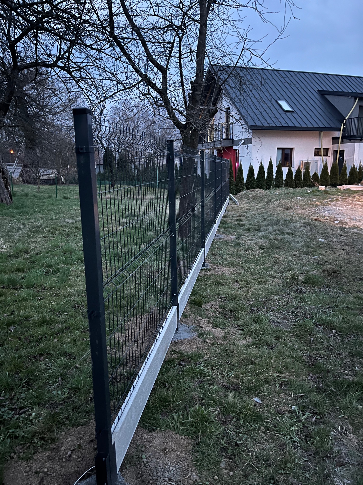 Nowoczesne ogrodzenie panelowe z podmurówką, zamontowane w Krakowie, na tle domu z ciemnym dachem i rzędem iglaków.