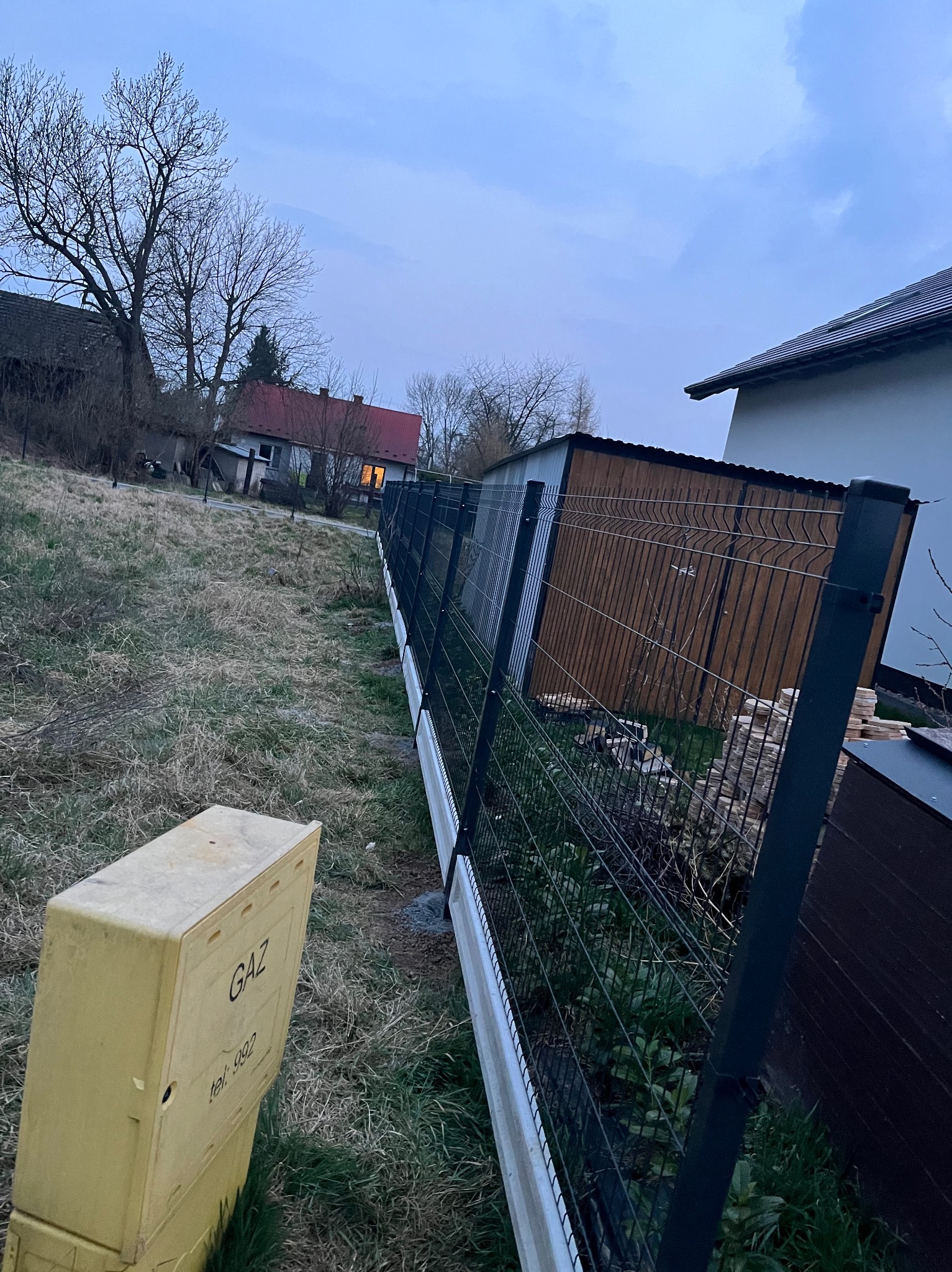 Nowoczesne ogrodzenie panelowe w kolorze grafitowym z podmurówką, widok z perspektywy skrzynki gazowej na tle zabudowań w pochmurny dzień.