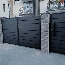 Nowoczesna, przesuwna brama w kolorze antracytowym z poziomymi panelami, osadzona na słupkach wykończonych szarą cegłą, na tle jasnej elewacji domu z balkonem.