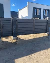 Nowoczesne, poziome ogrodzenie z szarych paneli kompozytowych na betonowej podmurówce, z furtką, na tle białego domu z ciemnymi oknami w słoneczny dzień.