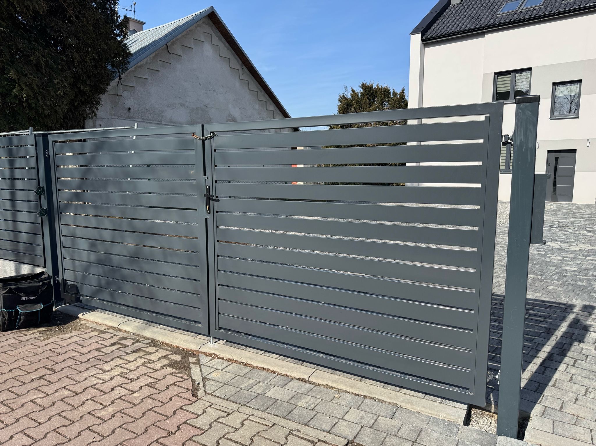 Nowoczesna, pozioma brama wjazdowa w kolorze grafitowym, z widocznym łańcuchem zabezpieczającym, na tle kostki brukowej i dwóch budynków o różnej architekturze.