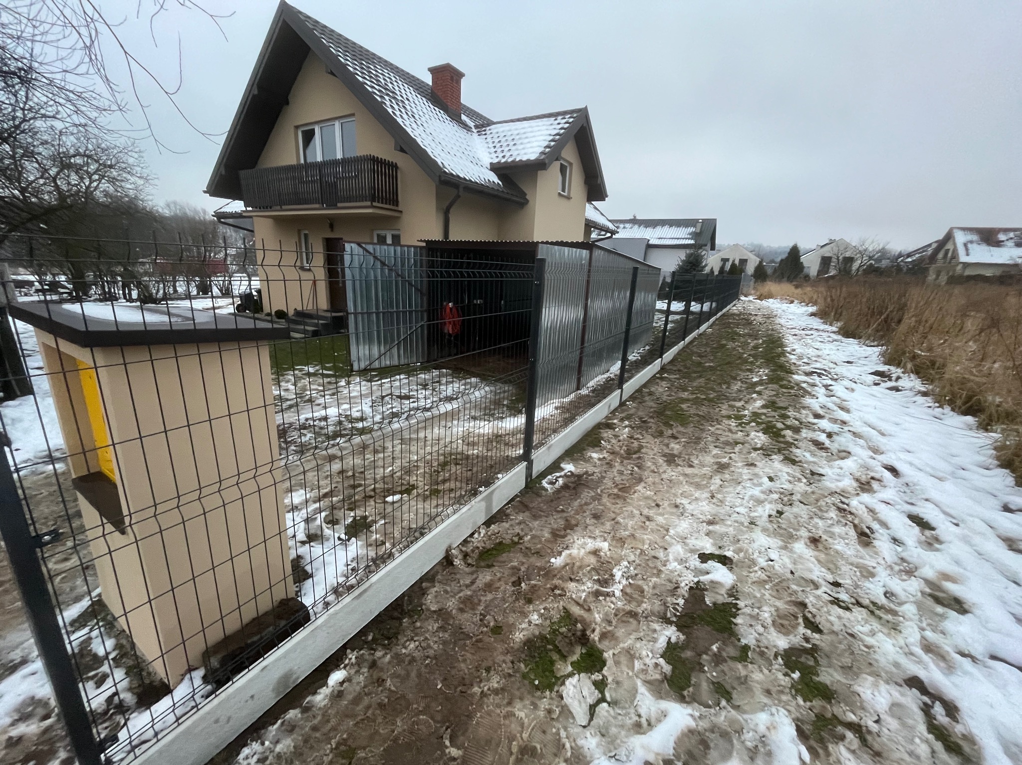 Nowoczesne ogrodzenie panelowe z podmurówką prefabrykowaną, przylegające do metalowej wiaty garażowej na tle domu w zimowej scenerii.