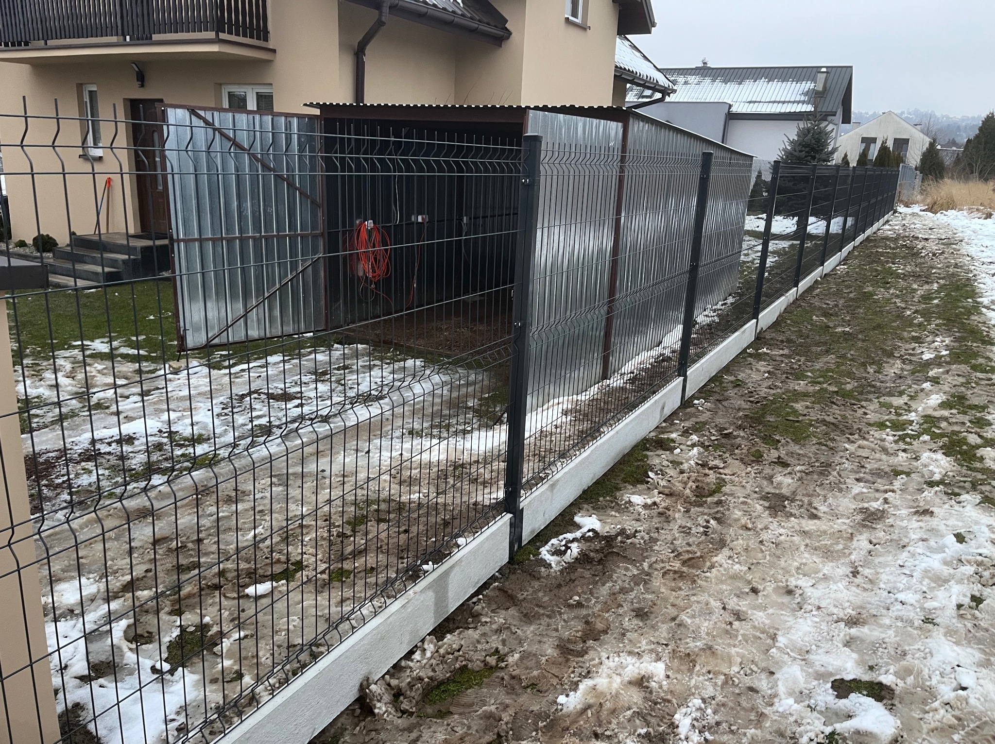 Nowoczesne ogrodzenie panelowe z podmurówką betonową, widok z zewnątrz, w tle budynek mieszkalny i garaż blaszany z otwartymi drzwiami, teren częściowo pokryty śniegiem.
