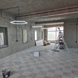 FERDI - Mateusz Major - Wnętrze w trakcie remontu: surowe ściany, szachownica płytek na podłodze, drabiny, nowoczesne lampy w kształcie sześciokątów. Widoczne ślady prac wykończeniowych.