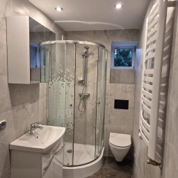 FERDI - Mateusz Major - Wykończona łazienka w Lubnie: kabina prysznicowa narożna z mozaiką, umywalka z szafką, grzejnik drabinkowy i toaleta. Szare płytki imitujące marmur na ścianach i podłodze.