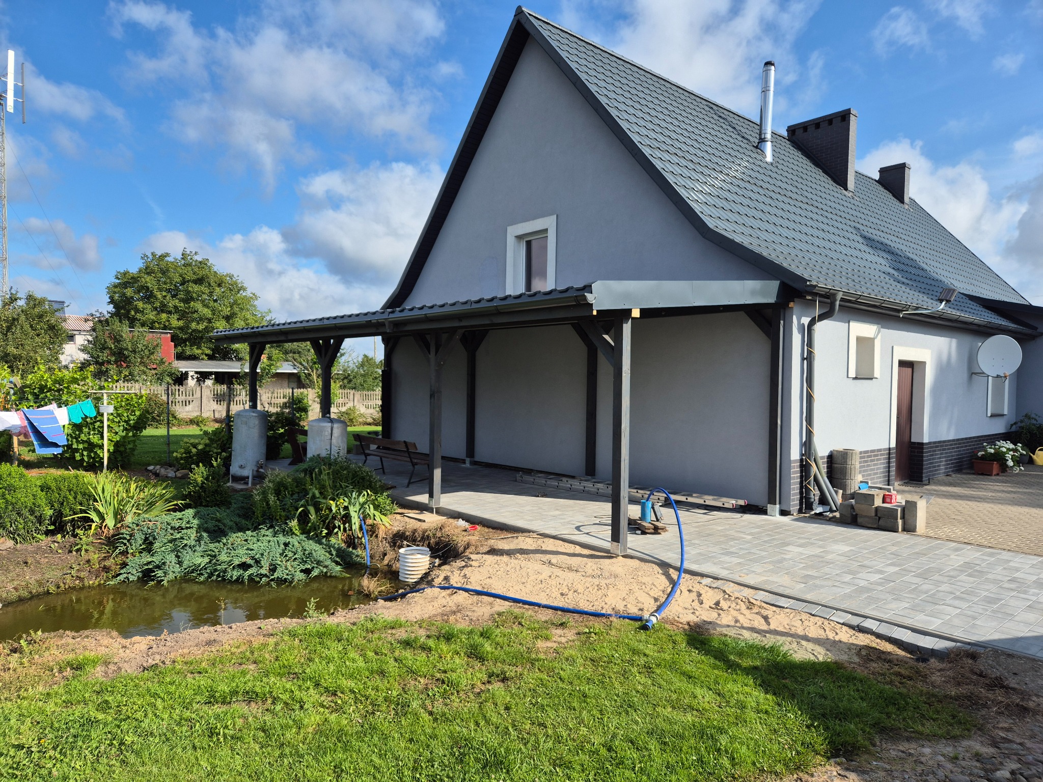 Nowoczesny dom z szarą elewacją i zadaszonym tarasem. Kostka brukowa ułożona na podjeździe, obok oczko wodne. Niebieski wąż ogrodowy na trawniku.