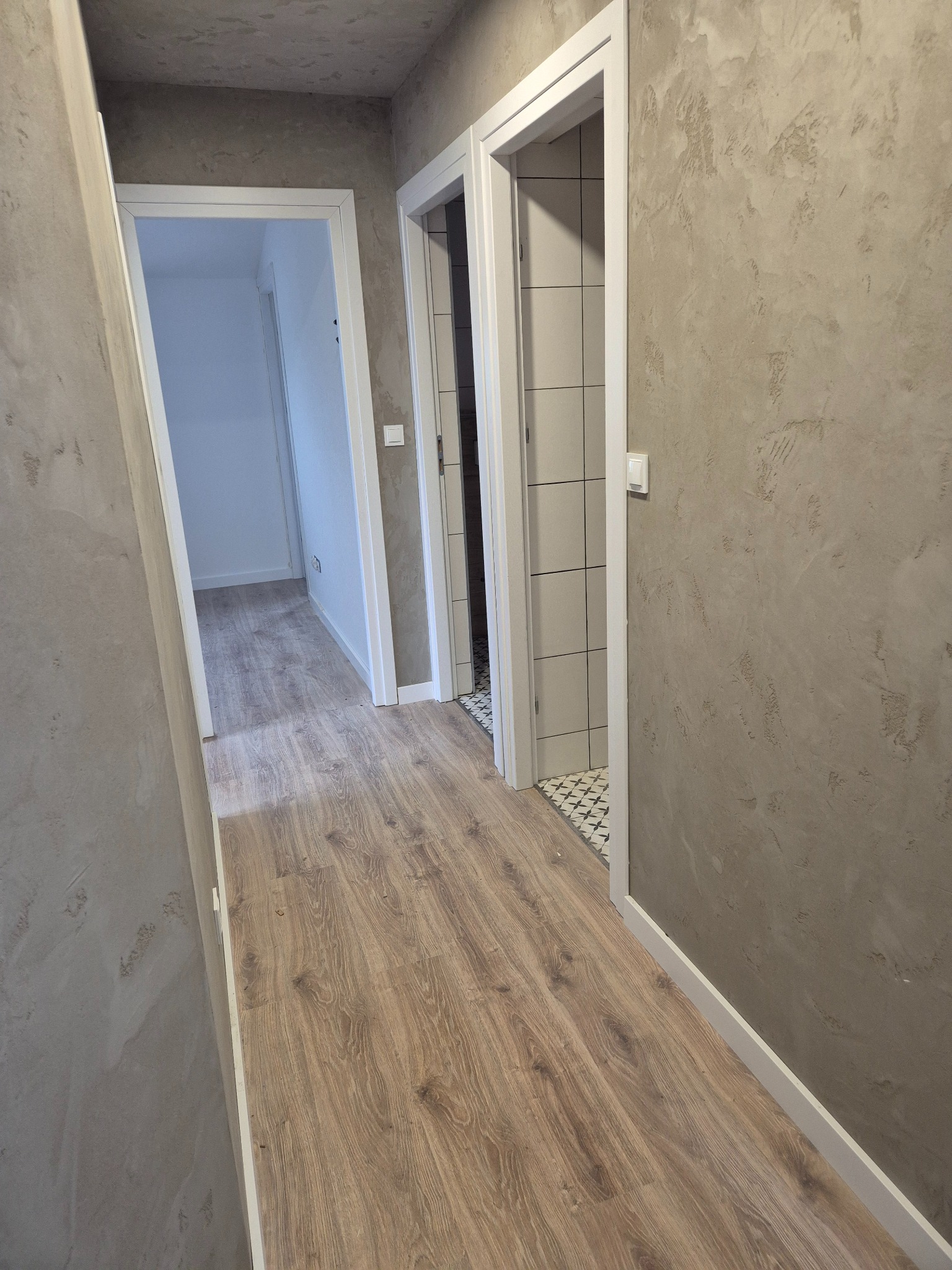 Korytarz po remoncie: panele podłogowe w odcieniach brązu, ściany pokryte dekoracyjnym tynkiem w beżowej tonacji, białe listwy przypodłogowe i ościeżnice.