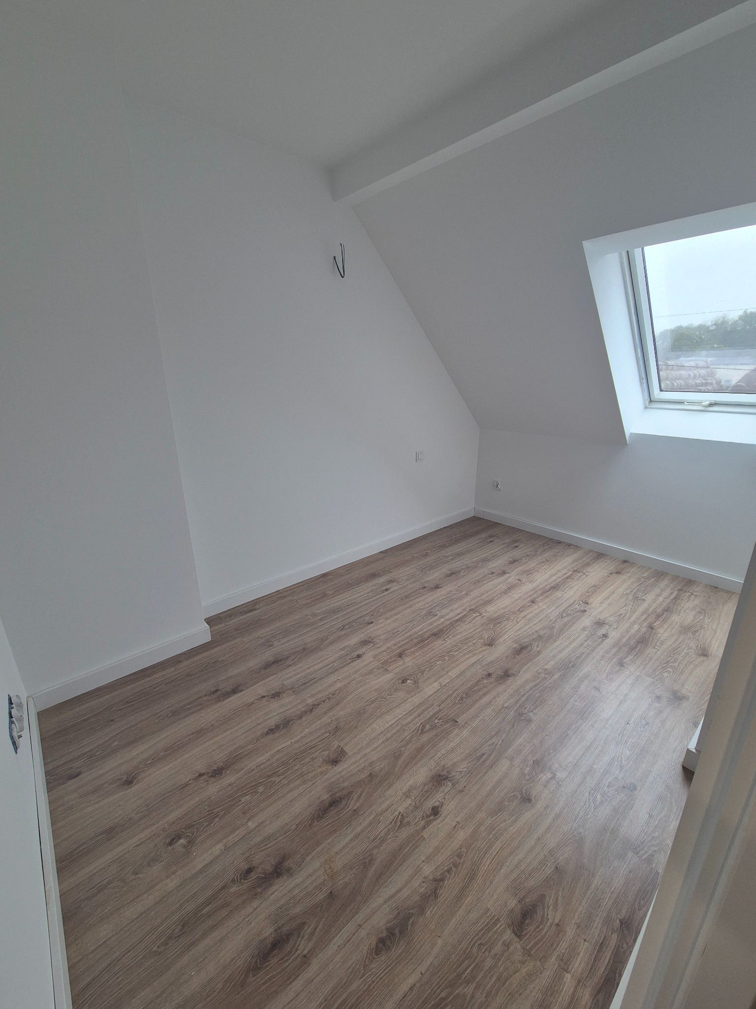 Jasne poddasze z oknem dachowym i panelami podłogowymi w odcieniach brązu. Ściany pomalowane na biało, widoczne listwy przypodłogowe. Minimalistyczny styl.