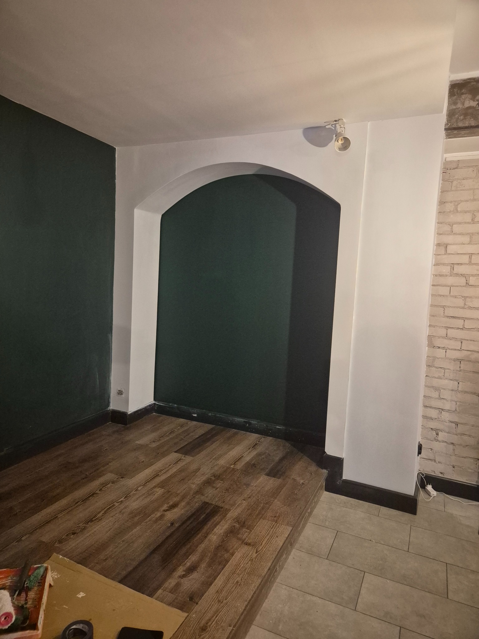 Wnętrze po remoncie: ciemnozielona ściana z łukowym przejściem, drewniana podłoga, fragment ściany z cegły. Widoczne narzędzia malarskie.