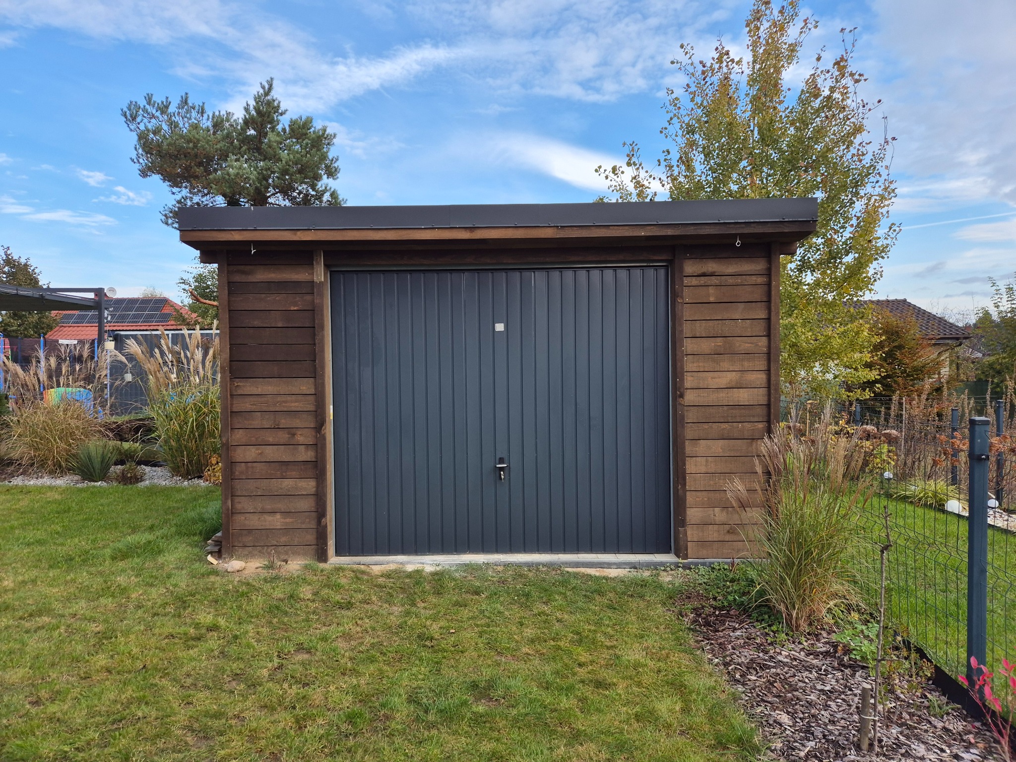 Nowoczesny, drewniany garaż z szarą bramą segmentową na tle zielonego trawnika i dekoracyjnych traw. Widoczne panele słoneczne na dachu domu w tle.