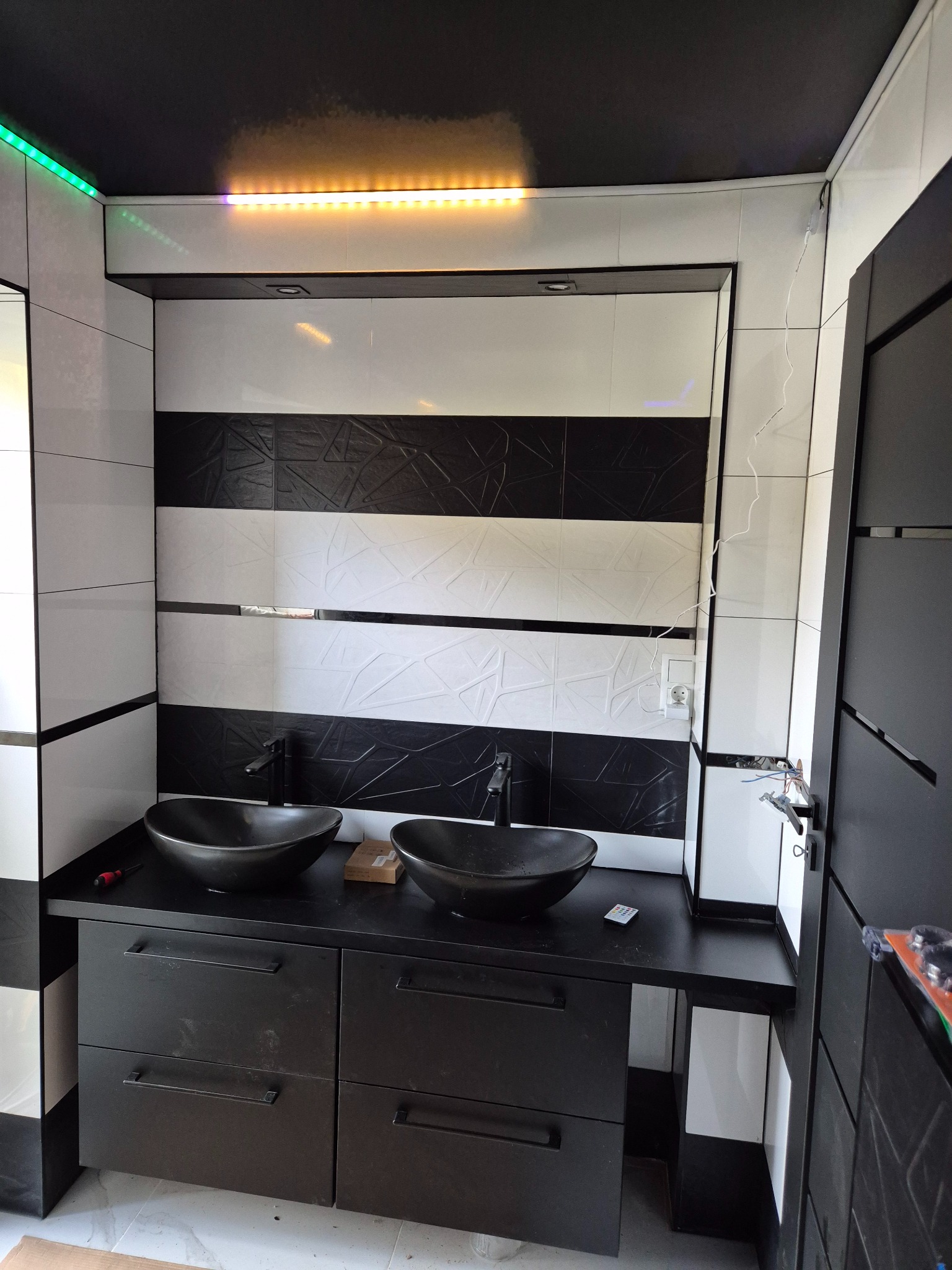 Wykończona łazienka z czarno-białymi płytkami, podwójną umywalką w kształcie misy i czarną szafką. Oświetlenie LED na suficie. Widoczne gniazdko elektryczne.