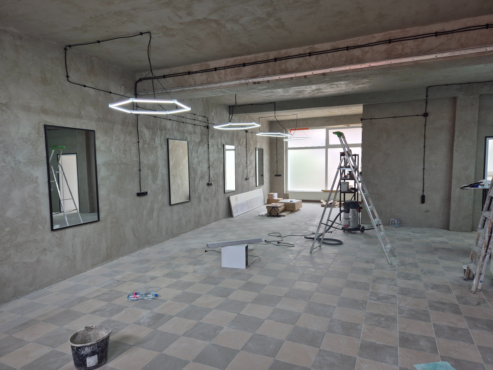 Wnętrze w trakcie remontu: surowe ściany, szachownica płytek na podłodze, drabiny, nowoczesne lampy w kształcie sześciokątów. Widoczne ślady prac wykończeniowych.