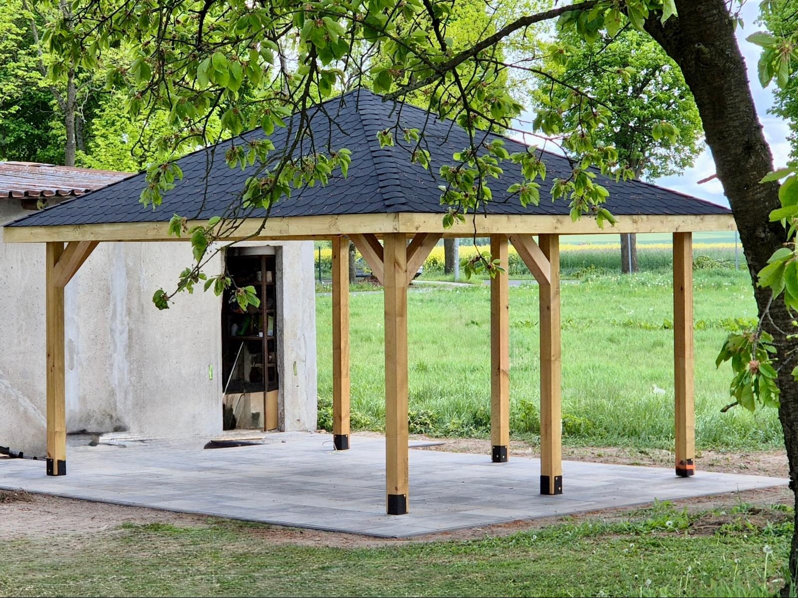 Drewniana altana z czarnym dachem z gontu bitumicznego, posadowiona na szarej kostce brukowej. Widok z zewnątrz, w tle zieleń i budynek gospodarczy.