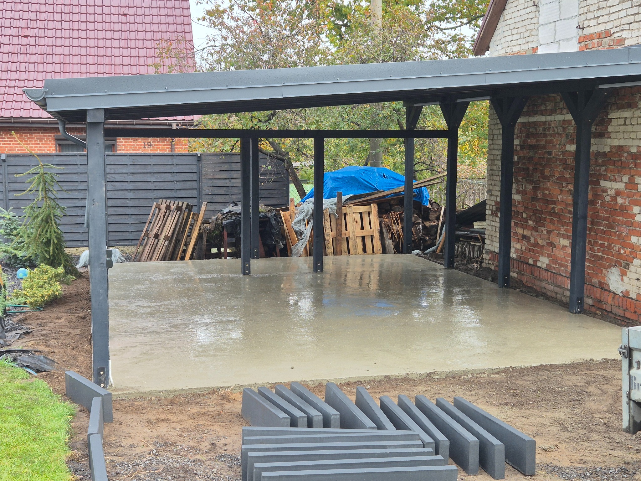 Świeżo wylana, mokra posadzka pod zadaszeniem z czarną konstrukcją. Na pierwszym planie ułożone szare obrzeża. W tle ceglany budynek i drewniane palety.