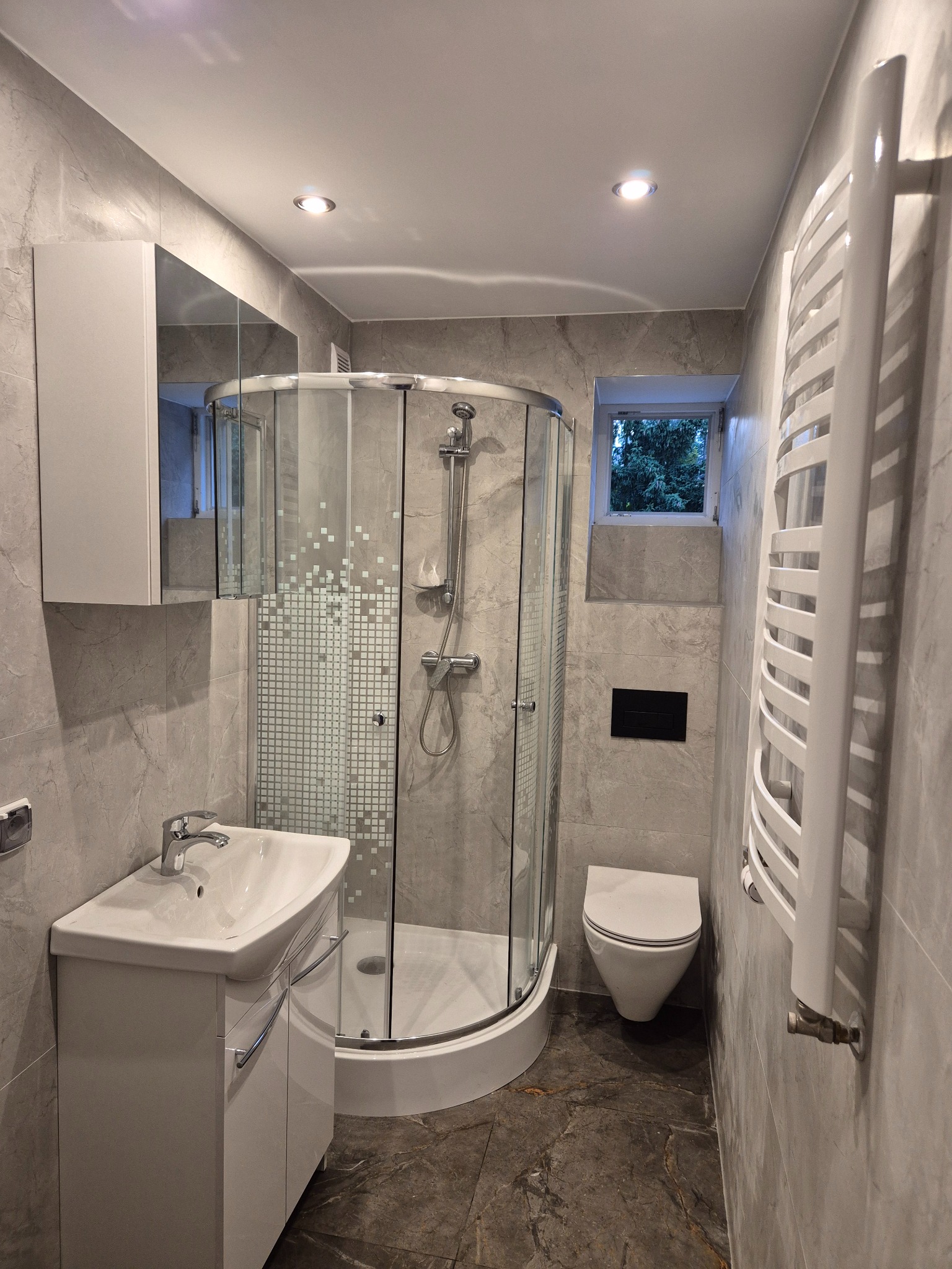 Wykończona łazienka w Lubnie: kabina prysznicowa narożna z mozaiką, umywalka z szafką, grzejnik drabinkowy i toaleta. Szare płytki imitujące marmur na ścianach i podłodze.