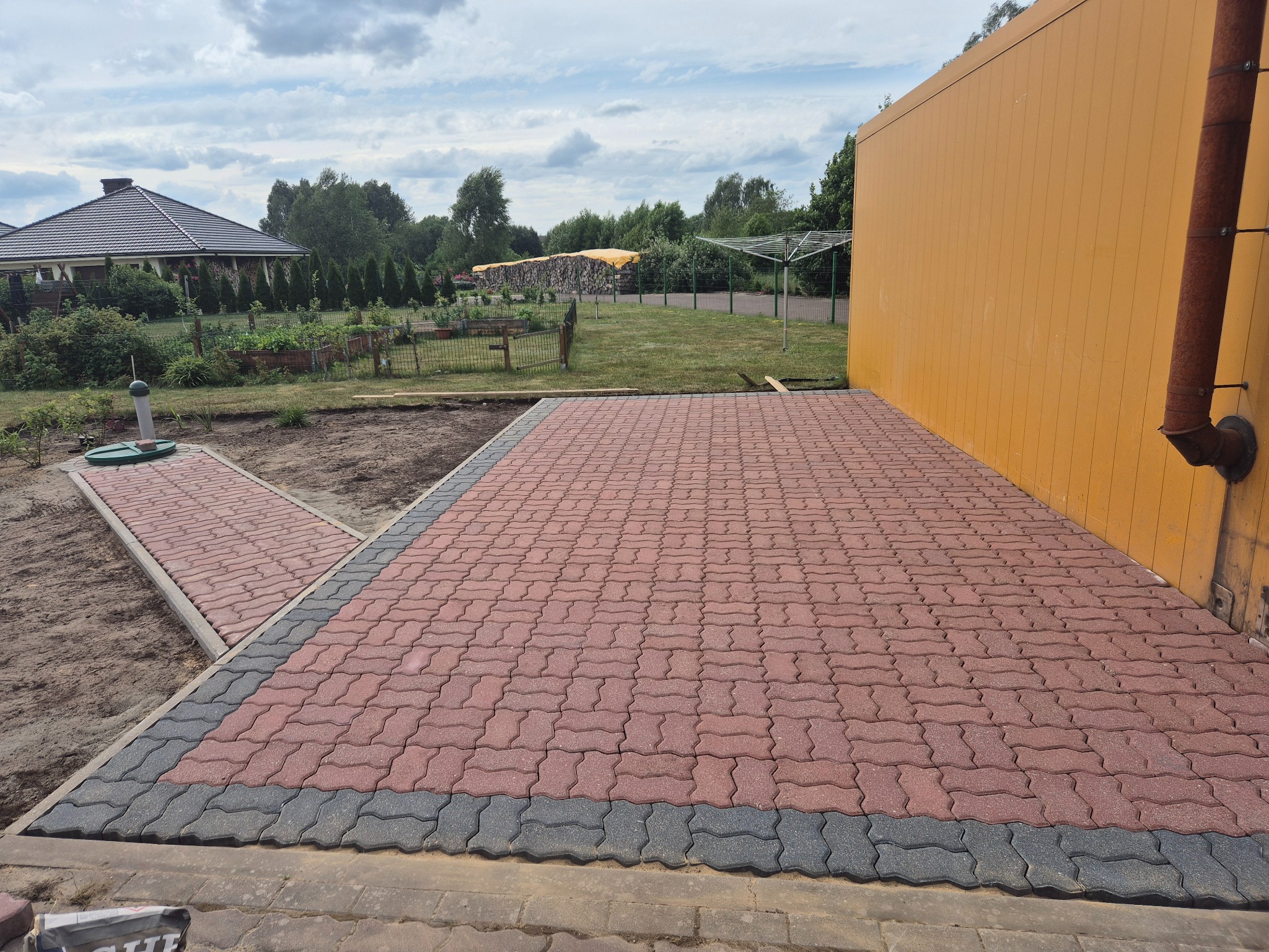 Ułożona czerwona kostka brukowa z szarym obramowaniem przy żółtej ścianie budynku. W tle widoczny dom i ogród pod pochmurnym niebem.