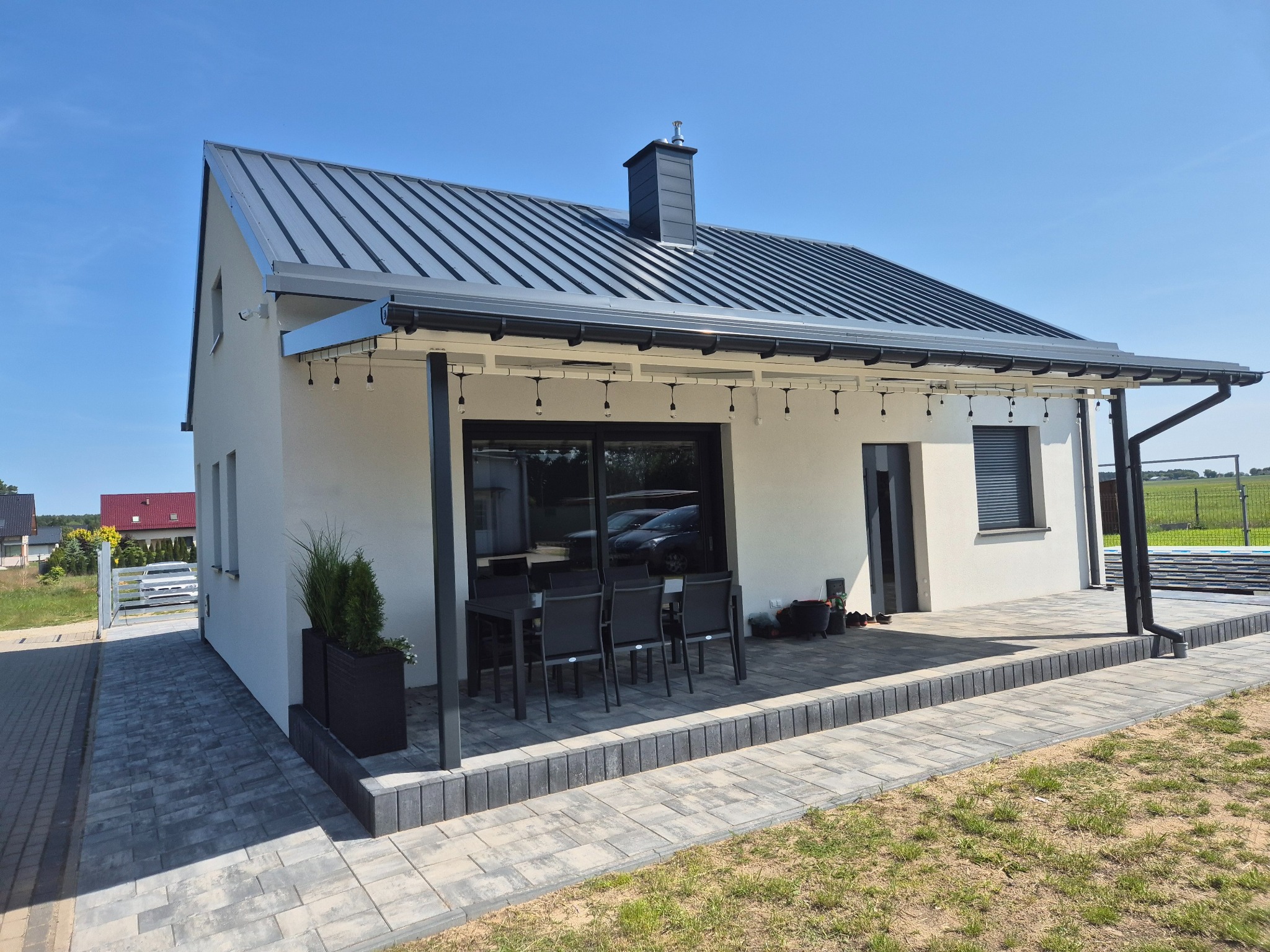 Nowoczesny dom jednorodzinny z szarą kostką brukową na tarasie i podjeździe, białą elewacją i ciemnym dachem z blachy na rąbek stojący w słoneczny dzień.