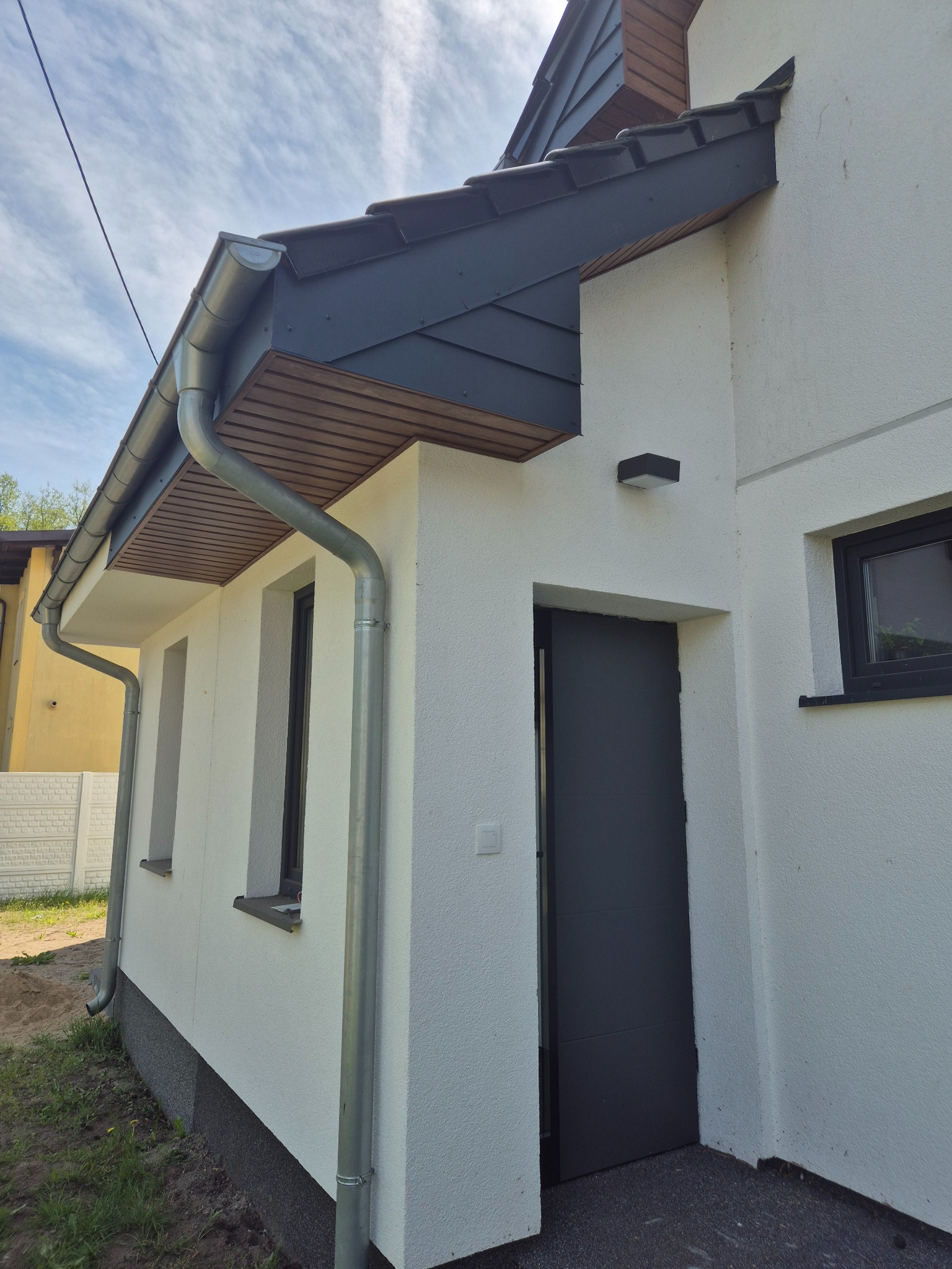 Elewacja domu z widocznymi oknami, drzwiami i orynnowaniem. Biała ściana z fakturą, ciemne okna i drzwi, srebrne rynny. Widok z dołu, błękitne niebo w tle.