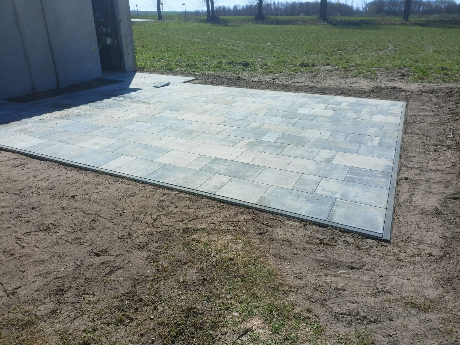 Ułożona, prostokątna nawierzchnia z szarych płyt betonowych, obramowana ciemną listwą. Widok na trawnik i budynek w tle. Świeżo ułożona kostka na ziemi.