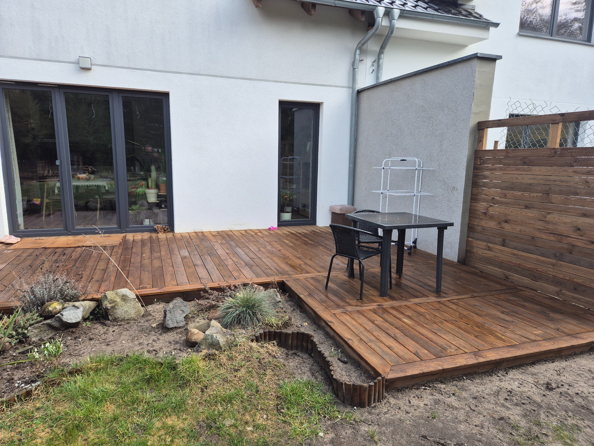 Drewniany taras przy domu z meblami ogrodowymi i rabatami kwiatowymi. Widoczne deski tarasowe, stół, krzesło i elementy dekoracyjne ogrodu.