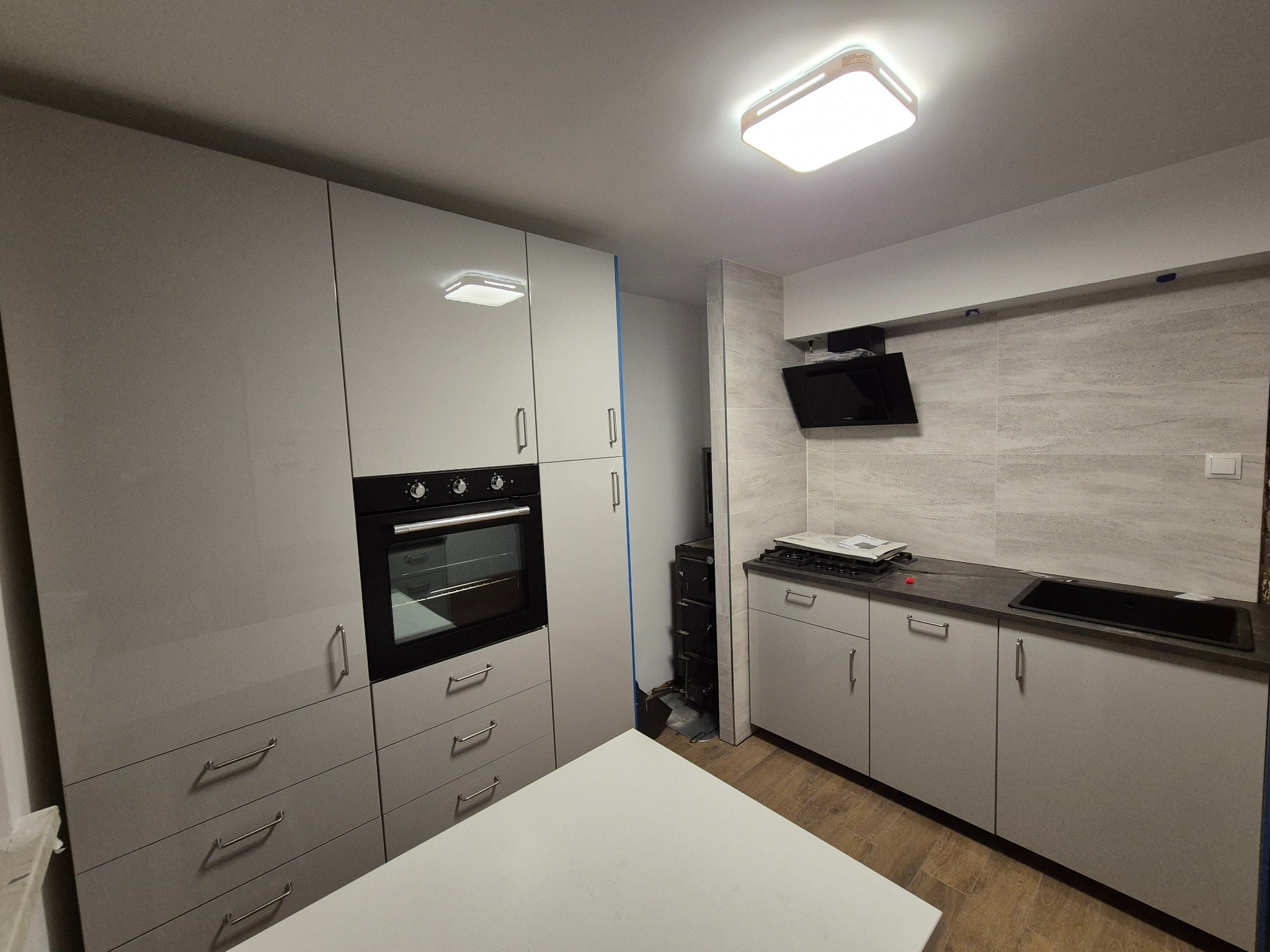 Jasna kuchnia z zabudową meblową, piekarnikiem i płytą grzewczą. Na ścianie płytki imitujące kamień. Oświetlenie sufitowe. Widok na fragment blatu roboczego.