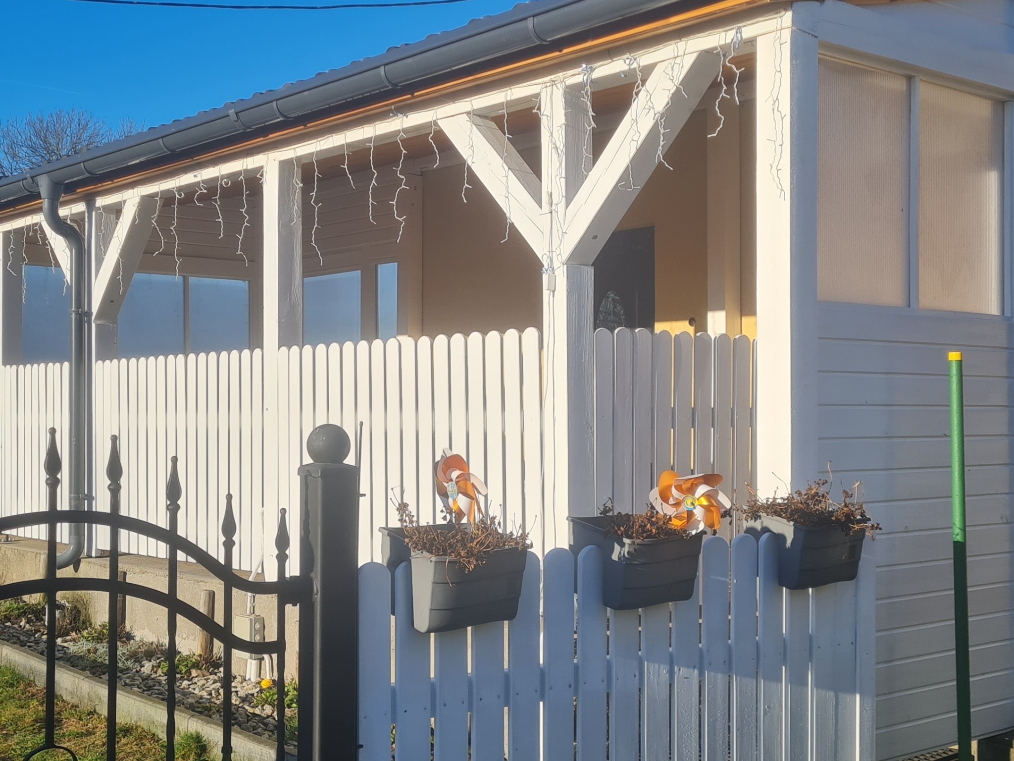 Biała, drewniana altana z dekoracjami i donicami na płocie, widziana zza czarnego, metalowego ogrodzenia w słoneczny dzień. Widoczne detale konstrukcyjne i elementy dekoracyjne.