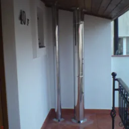 Dwa chromowane kominy odprowadzające spaliny, wystające z podłogi balkonu z brązowych płytek, z drewnianym sufitem i fragmentem metalowej balustrady.