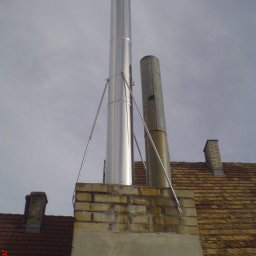 ZAKŁAD URZĄDZEŃ GRZEJNYCH I ŚLUSARSTWA ANDRZEJ KREFT w spadku - Nowy, błyszczący komin ze stali nierdzewnej zamontowany obok starego, skorodowanego komina na ceglanym dachu z widocznymi mocowaniami.